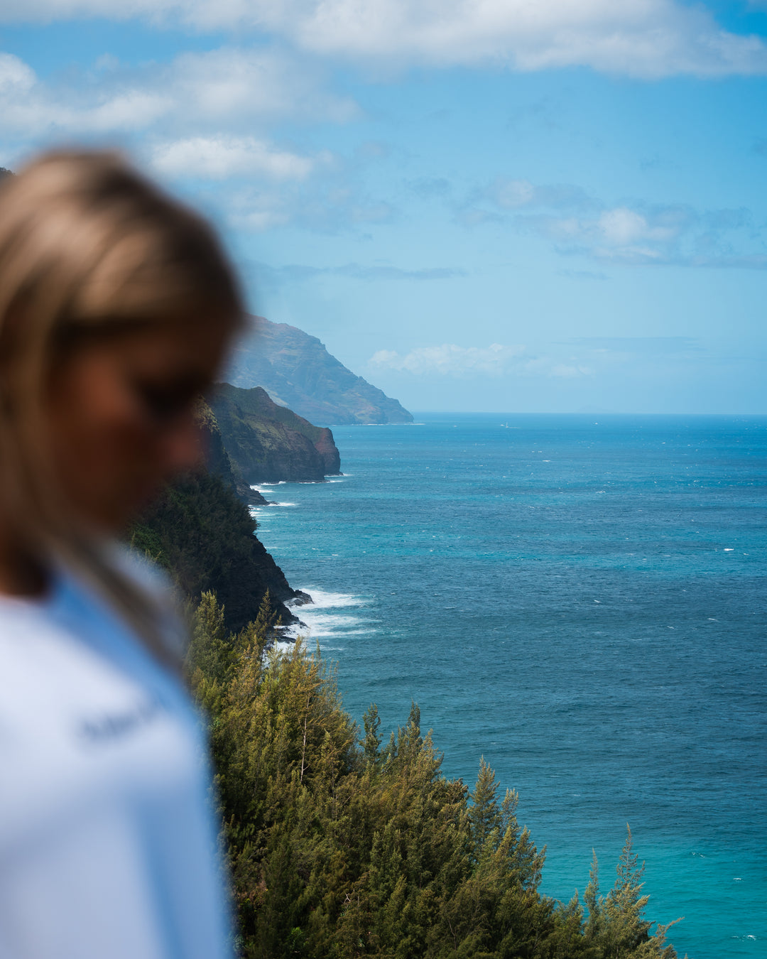 A scenic coastal landscape with a deep blue ocean, green cliffs, and a partly cloudy sky, with a blurred person in the foreground on the left.