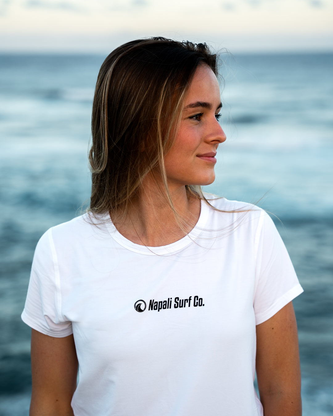 A woman with long brown hair stands by the ocean wearing a white short-sleeve modal surf t-shirt with the black logo Napali Surf Co. printed on the chest.
