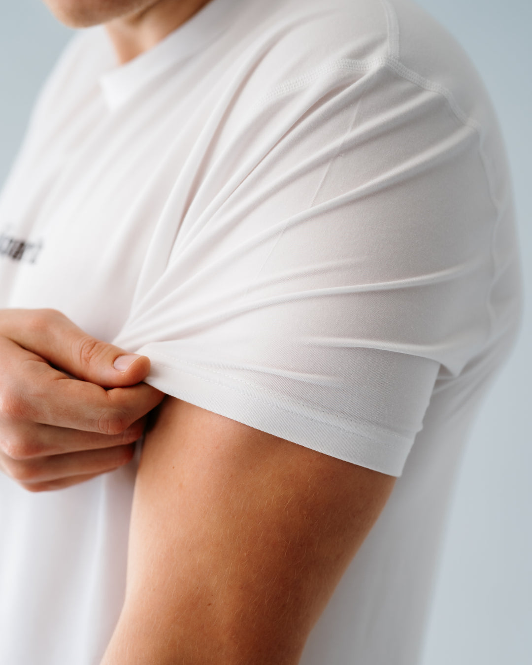 Close-up of a person wearing a white short-sleeve modal surf T-shirt, pulling the sleeve upward to show the fabric stretch and fit against a light gray background.